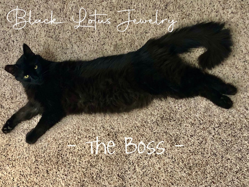 May include: A black cat with yellow eyes is lying on a carpet. The text "Black Lotus Jewelry" is written above the cat, and the text "- The Boss -" is written below the cat.