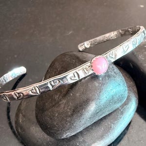 May include: A silver bangle bracelet with a pink stone and heart-shaped engravings.