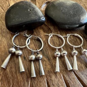 May include: Two pairs of silver hoop earrings, each featuring two dangling charms consisting of a small sphere attached to a slender cone. The earrings are crafted from polished silver metal, showcasing a circular hoop design with a secure wire closure.