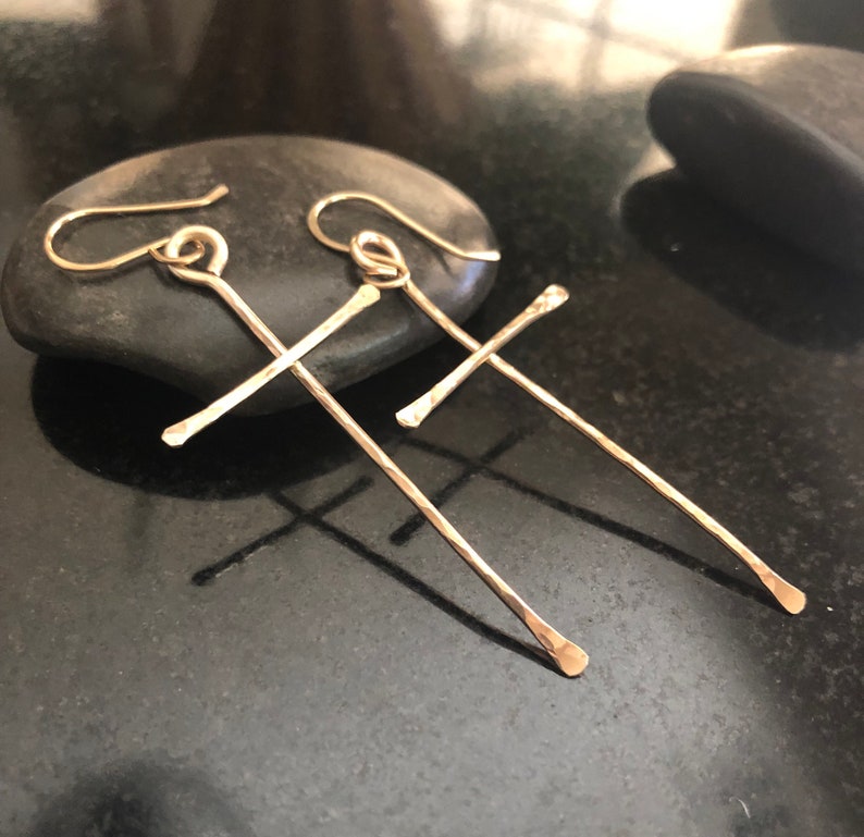 May include: A pair of gold-colored cross earrings with hammered texture. The earrings are displayed on a black surface with a dark stone in the background. The earrings have a hook closure.