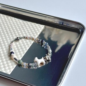 May include: A beaded bracelet with a white bone-shaped charm. The bracelet features a mix of colorful beads in shades of pink, blue, green, and clear. It is displayed on a silver tray with a textured white surface.