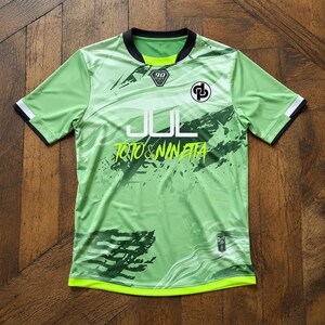 May include: A light green short-sleeve jersey with a black crew neck collar and sleeve cuffs. The front features the text JUL and TOTO & NINETTA in white and lime green, a black circular logo, a 90 emblem, and dark green abstract graphic patterns.