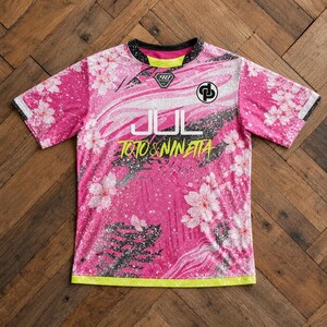 May include: A short-sleeve crew neck shirt in vibrant pink featuring a white cherry blossom pattern, black paint splatter accents, and a neon yellow hem. The chest displays the white text JUL, yellow text TOTO & NINETTA, and two logos.