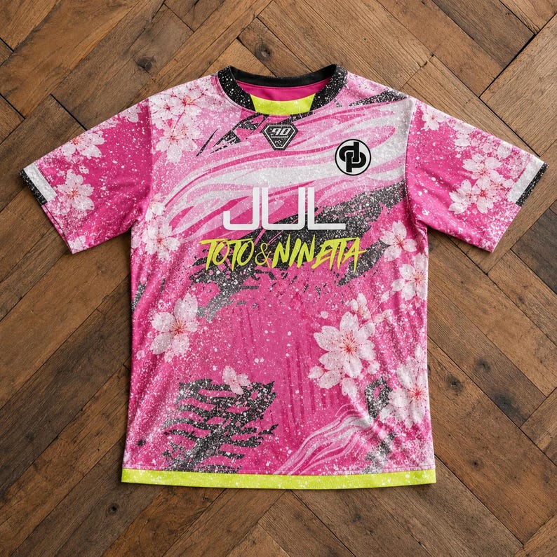 May include: A short-sleeve crew neck shirt in vibrant pink featuring a white cherry blossom pattern, black paint splatter accents, and a neon yellow hem. The chest displays the white text JUL, yellow text TOTO & NINETTA, and two logos.