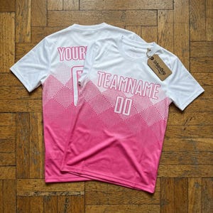 Peut inclure: Deux t-shirts de sport blancs avec un dégradé rose. L'un porte l'inscription "YOUR" et l'autre "TEAMNAME 00". Les deux t-shirts ont une étiquette "UrbanDrip".