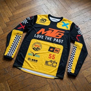 Può includere: Maglia a maniche lunghe nera e gialla con la scritta "MTB LOVE THE PAST" e il numero "55". La maglia presenta vari loghi e motivi a scacchi sulle maniche e sulle spalle.