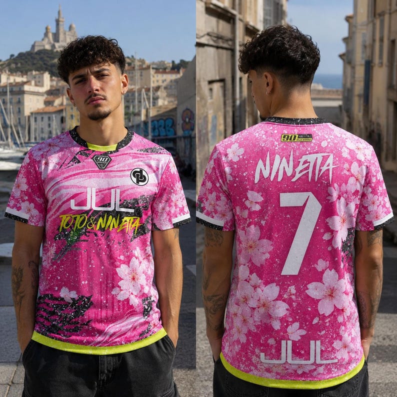 May include: A short-sleeved athletic jersey in vibrant pink featuring a white cherry blossom pattern, black splatter accents, and a neon yellow hem. The front displays the text JUL and TOTO & NINETA, while the back features the name NINETA and number 7.