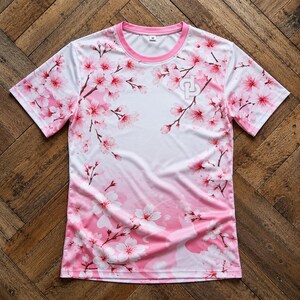 May include: A short-sleeve t-shirt featuring a white base with a pink cherry blossom floral pattern. The shirt has a pink crew neckline and a small white logo on the upper right chest. The design transitions into a solid pink gradient at the hem.