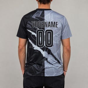 May include: A short-sleeve t-shirt featuring a split design with a black left side and a gray right side. The back displays the text YOURNAME above the number 00 in a white font with a black outline. A jagged white diagonal graphic separates the colors.