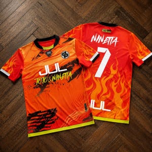 May include: Two short-sleeve jerseys featuring a vibrant orange and red flame pattern. The front displays the text JUL and TOTO & NINETTA with a circular logo. The back features the name NINETTA, the number 7, and the text JUL with a small 90 logo.