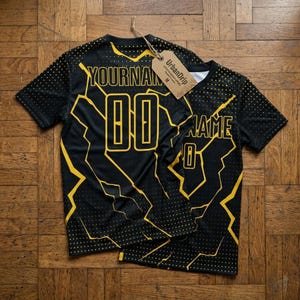 Peut inclure: Deux t-shirts de sport noirs avec des motifs d'éclairs jaunes et les textes "YOURNAME" et "NAME" en jaune. Les chiffres "00" et "0" sont également en jaune. Les chemises ont une étiquette "UrbanDrip".