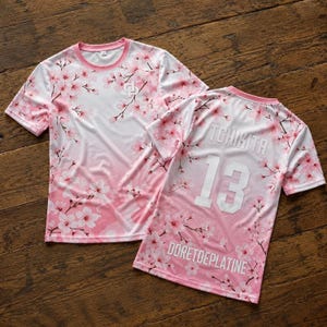 May include: Two short-sleeve t-shirts featuring a pink and white cherry blossom pattern. The front shirt has a small logo on the chest. The back shirt displays the text TCHIKITA, the number 13, and the text DORETDEPLATINE in white lettering.