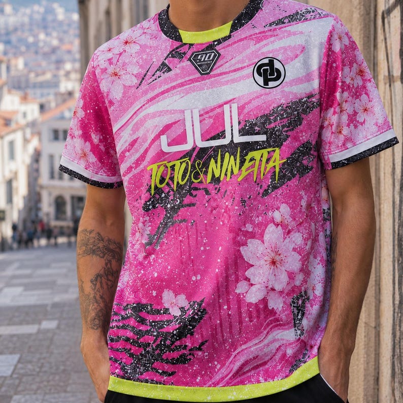 May include: A short-sleeve t-shirt featuring a vibrant pink and white cherry blossom pattern with black splatter accents. The front displays the text JUL and TOTO & NINETTA in yellow, along with a circular logo and a diamond-shaped 90 emblem.