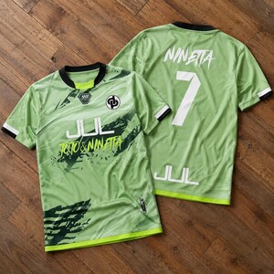 May include: Two light green short-sleeve jerseys with black trim. The front features a black logo, the text JUL, and TOTO & NINETTA. The back displays the name NINETTA, the number 7, and the text JUL in white. Both shirts have a dark green graphic pattern.