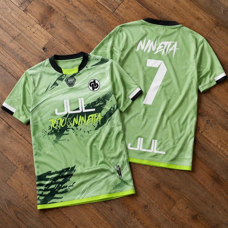 May include: Two light green short-sleeve jerseys with black trim. The front features a black logo, the text JUL, and TOTO & NINETTA. The back displays the name NINETTA, the number 7, and the text JUL in white. Both shirts have a dark green graphic pattern.