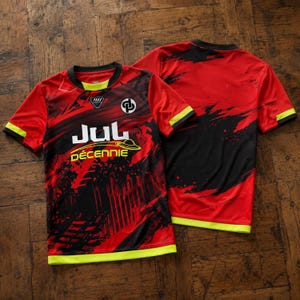 May include: A short-sleeved athletic jersey featuring a red and black abstract brushstroke pattern. The front displays the text JUL D&Eacute;CENNIE with a yellow graphic, a circular logo, and a small black emblem. The collar and hem have neon yellow trim.