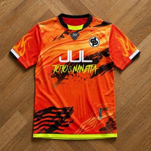 May include: A short-sleeve athletic jersey in vibrant orange and red with a flame pattern on the sleeves. It features the text JUL and TOTO & NINETTA in white and yellow, a black circular logo, a 90 emblem at the collar, and a neon yellow hem.