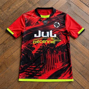 May include: A short-sleeved t-shirt featuring a red and black abstract graphic pattern. The shirt has a black crew neckline with a lime green accent, lime green sleeve cuffs, and a lime green hem. It displays the text JUL and D&Eacute;CENNIE.