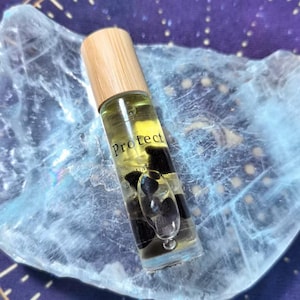 Puede incluir: Un frasco de aceite esencial en rollo de vidrio transparente con tapa de madera. El frasco contiene un líquido amarillo y piedras negras. La palabra "Protect" está impresa en el frasco. El frasco descansa sobre una superficie azul y blanca.