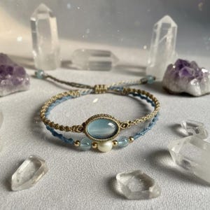 May include: A double-strand bracelet featuring a central oval light blue cabochon set in tan macrame cord. The second strand consists of light blue cord accented with a single white pearl, two small light blue beads, and two tiny gold-toned metal beads.