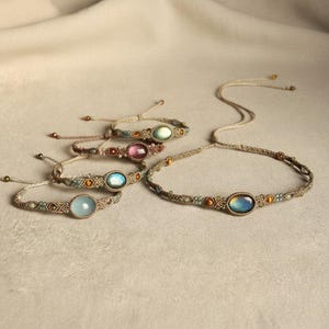 May include: A collection of handmade macrame jewelry, including bracelets and a necklace. Each piece features a central oval gemstone in various colors, such as blue, pink, and green, and is accented with small beads. The jewelry is displayed on a neutral surface.