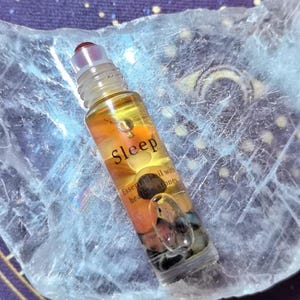 May include: A clear glass essential oil roller bottle with the word "Sleep" printed on it. The bottle contains a yellow liquid and small stones. The bottle has a red cap and is resting on a light blue surface.