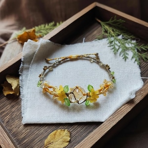 Op de afbeelding: Een handgemaakte armband met een centraal helder kristal, versierd met gele bloemenaccenten en groene bladeren. De armband is gemaakt met een geweven koord en kleine kralen, geplaatst op een witte doek in een houten dienblad.