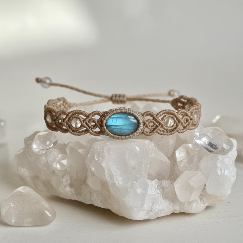 May include: A macrame bracelet with a central oval-shaped blue gemstone. The bracelet is made of tan-colored cord with a woven design. Clear beads are incorporated into the design. The bracelet is displayed on a white, crystalline surface.