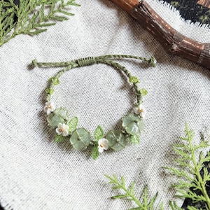 Könnte beinhalten: Ein handgefertigtes grünes Armband mit hellgrünen Edelsteinperlen, kleinen weißen Blüten und grünen Blättern. Das Armband ist an einem grünen Kordelband befestigt und wird auf einer strukturierten, neutralfarbenen Oberfläche präsentiert.