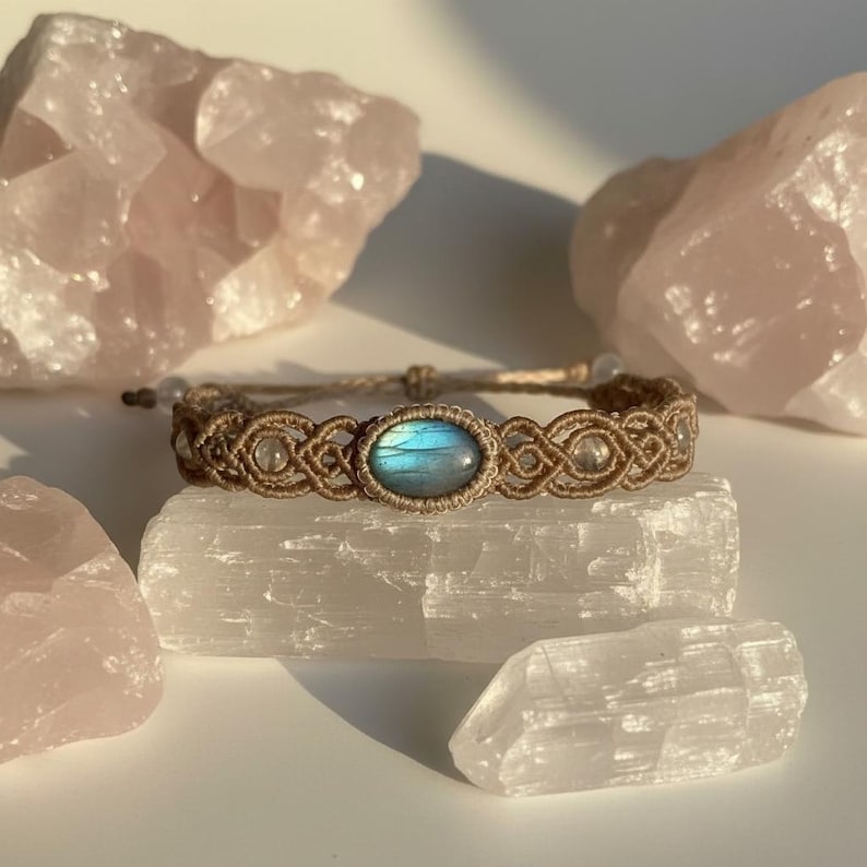 May include: A handcrafted macrame bracelet featuring a central oval-shaped blue gemstone. The bracelet is made with tan-colored cord and accented with clear beads. The bracelet is displayed on a white crystal.