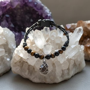 Może przedstawiać: Czarna i beżowa bransoletka z koralik&oacute;w z tygrysim okiem i czarnych koralik&oacute;w. Z centralnej części wisi owinięty drutem, ciemnofioletowy kryształowy wisiorek. Bransoletka jest prezentowana na kryształach kwarcu.