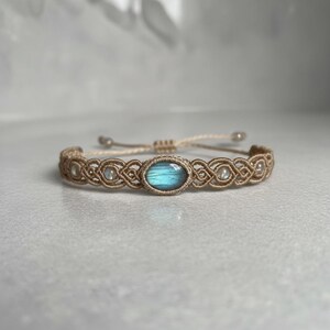 May include: A macrame bracelet with a central oval-shaped blue gemstone and smaller white beads. The bracelet is made of tan-colored cord and has an adjustable sliding knot closure. The design features intricate knotting patterns.