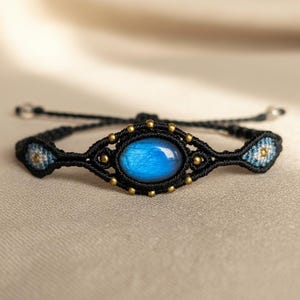 May include: A handmade macrame bracelet featuring a vibrant blue gemstone center. The black cord is accented with gold-colored beads and small, patterned details in blue and white. The bracelet is designed with an adjustable closure.