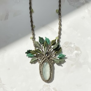 May include: A handmade macrame necklace featuring a tree design. The pendant has a light green oval stone at the center, surrounded by a macrame tree with green leaves. The necklace cord is a light brown color with small green beads.