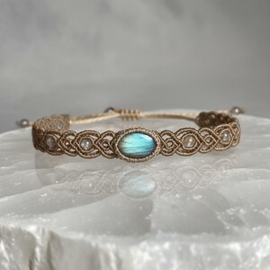 May include: A handmade macrame bracelet featuring a central oval-shaped, blue-green gemstone. The bracelet is crafted with tan-colored cord, showcasing intricate knotting patterns. Small, clear beads are interspersed along the design, adding subtle detail to this piece of jewelry.