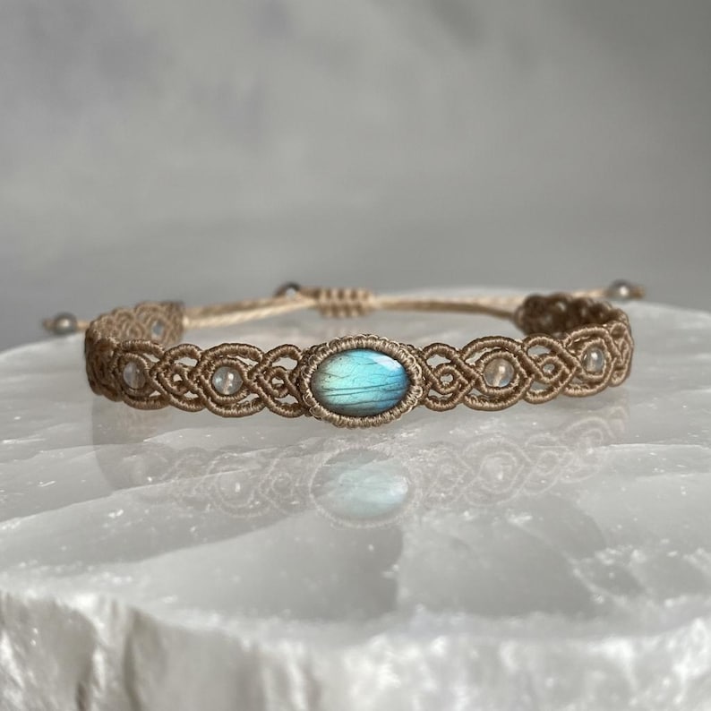 May include: A handmade macrame bracelet featuring a central oval-shaped, blue-green gemstone. The bracelet is crafted with tan-colored cord, showcasing intricate knotting patterns. Small, clear beads are interspersed along the design, adding subtle detail to this piece of jewelry.