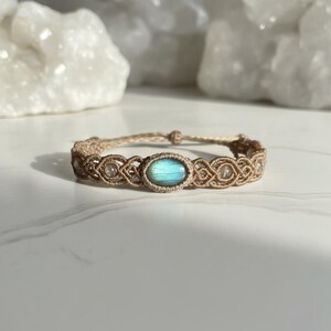 May include: A macrame bracelet featuring a central oval-shaped, iridescent blue-green gemstone. The bracelet is crafted with tan-colored cord and includes small, clear beads. The design is intricate and the bracelet is displayed on a white surface.