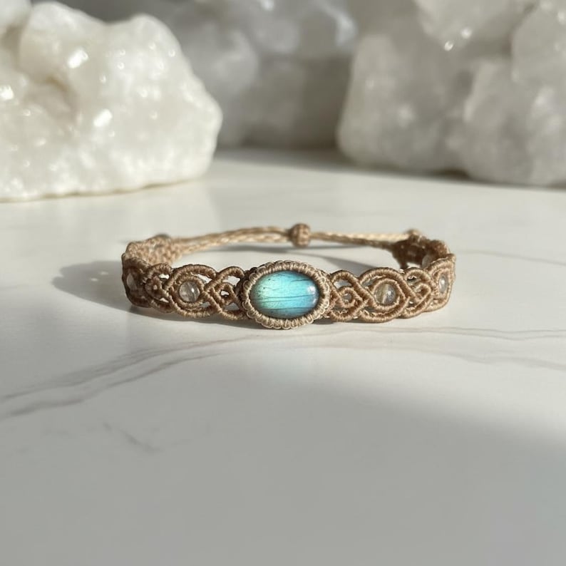 May include: A macrame bracelet featuring a central oval-shaped, iridescent blue-green gemstone. The bracelet is crafted with tan-colored cord and includes small, clear beads. The design is intricate and the bracelet is displayed on a white surface.