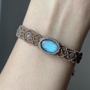 May include: A handmade macrame bracelet featuring a central oval-shaped blue gemstone, flanked by smaller pink stones. The bracelet is crafted with tan-colored cord, showcasing intricate knotting patterns. A delicate piece of jewelry.