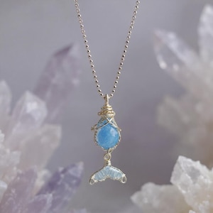 May include: A silver-toned necklace featuring a light blue, wire-wrapped pendant with a round bead and a tail-shaped charm. The necklace has a delicate chain and a whimsical, handcrafted aesthetic.
