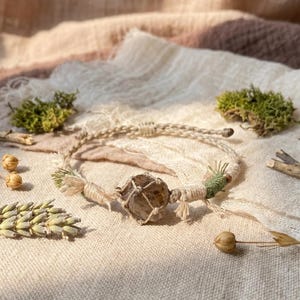 May include: A macrame bracelet featuring a central raw smoky quartz crystal encased in light beige cord netting. The bracelet band is crafted from braided beige cord with decorative knots and a small section of green thread accents.