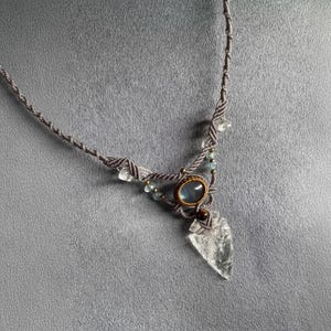 May include: A handmade macramé necklace with a central oval blue gemstone, surrounded by smaller blue and clear crystal accents. The pendant includes a clear, arrowhead-shaped crystal. The necklace is made with grey cord.