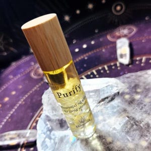 May include: A clear glass essential oil roller bottle with a wooden cap. The bottle contains a yellow liquid and small stones. The text "Purify" is printed on the bottle, along with "Essential oil with healing stones" and the brand name "LUMINAZ".