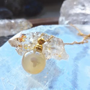 Może przedstawiać: Złoty naszyjnik z jasnożółtą, kulistą zawieszką. Zawieszka ma złotą nasadkę. Łańcuszek naszyjnika jest również złoty. Zawieszka spoczywa na przezroczystej, krystalicznej powierzchni. To biżuteria.