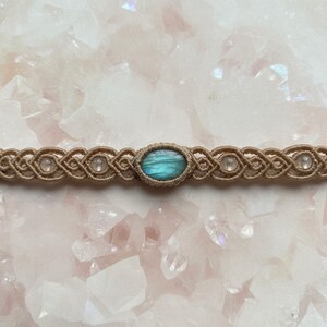 May include: A handcrafted macrame choker necklace featuring a central oval labradorite stone with blue and green hues. The beige cord is woven with intricate knots, accented by small, clear beads. The necklace is displayed on a pink surface.