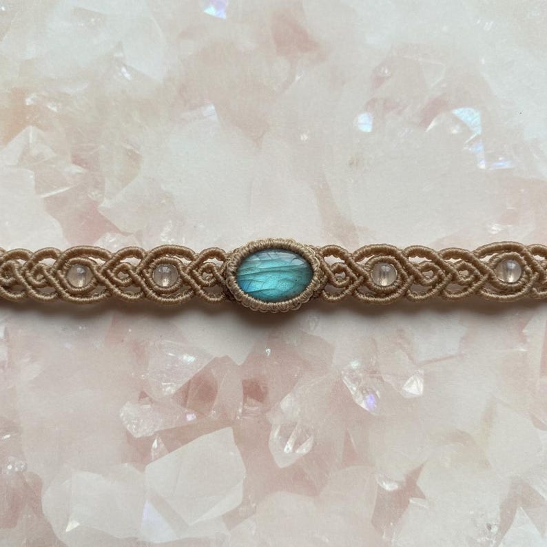 May include: A handcrafted macrame choker necklace featuring a central oval labradorite stone with blue and green hues. The beige cord is woven with intricate knots, accented by small, clear beads. The necklace is displayed on a pink surface.