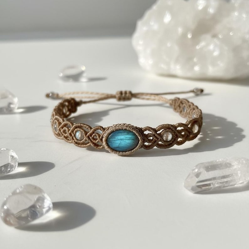 May include: A macrame bracelet with a central oval blue gemstone and clear crystal accents. The bracelet is made of tan-colored cord and has an adjustable sliding knot closure. Additional clear crystals are scattered around the bracelet.