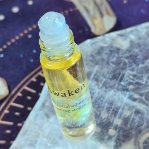 May include: A clear glass essential oil roller bottle with a light blue cap. The bottle contains a yellow liquid and has the word "Awaken" printed on it. The text "essential oil with healing stones" is also visible.