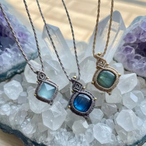 May include: Three macrame necklaces with gemstone pendants. The pendants are various shapes and colors, including light blue, dark blue, and green. The necklaces have a neutral-colored cord and small gold accents. The background features crystals and a purple geode.