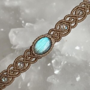 May include: A handmade macrame bracelet featuring a central oval labradorite stone with blue and green iridescence. The bracelet is woven with tan-colored cord and includes small, clear beads. The intricate knotwork creates a decorative pattern.